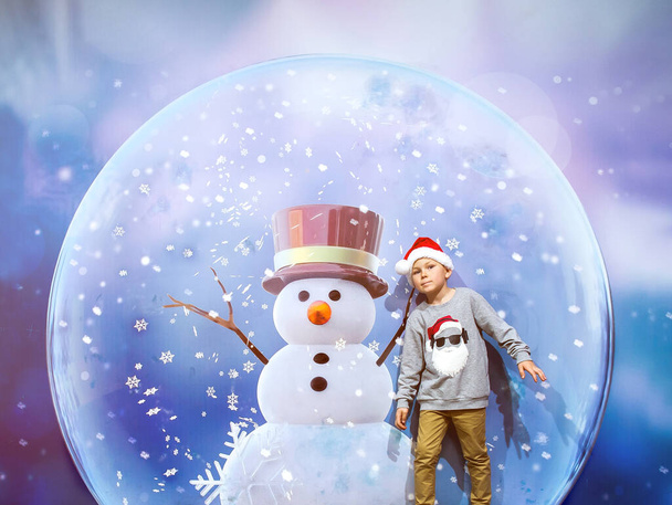 Счастливый мальчик и снеговик в волшебном шаре. Удивлённый ребёнок внутри рождественского снежного шара. Маленький Санта Клаус под снежинками. С Новым годом и Рождеством. Мальчик в волшебном месте на праздники. - Фото, изображение