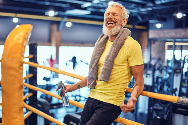 Счастливый зрелый мужчина с полотенцем на плечах, смотрящий в камеру и улыбающийся, стоящий на ринге и отдыхающий после тренировки по боксу в спортзале - Фото, изображение