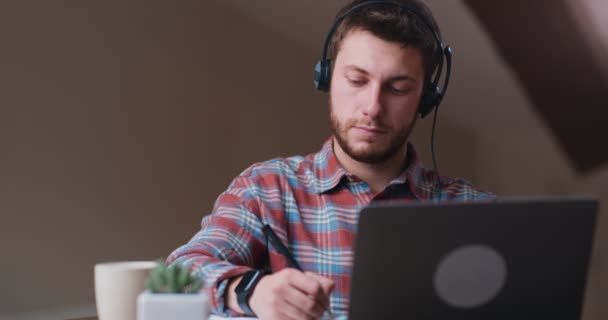 Nuevo aprendizaje normal y remoto. Hombre joven en auriculares viendo webinar en línea y tomando notas en casa - Metraje, vídeo