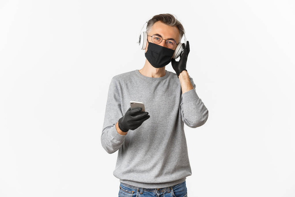 Konzept des covid-19, soziale Distanzierung und Lebensstil. Bild eines Mannes mittleren Alters mit medizinischer Maske, Handschuhen und Brille, Musik über Kopfhörer hörend und Smartphone in der Hand - Foto, Bild