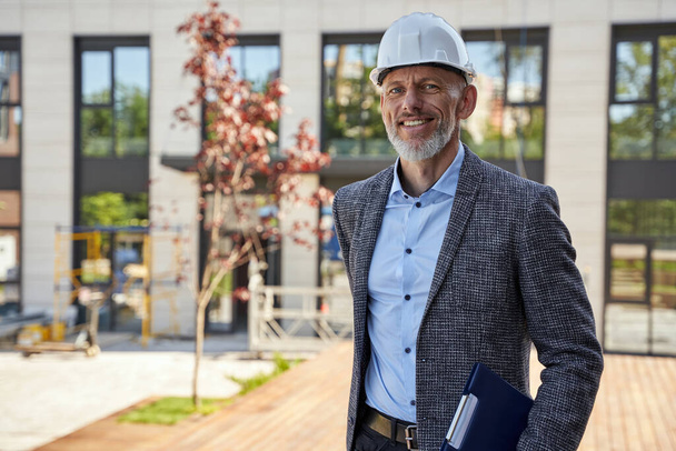 Modern architect. Successful middle aged business man, engineer manager wearing helmet looking at camera, standing outdoors and holding a blueprint while planning new project - Photo, Image