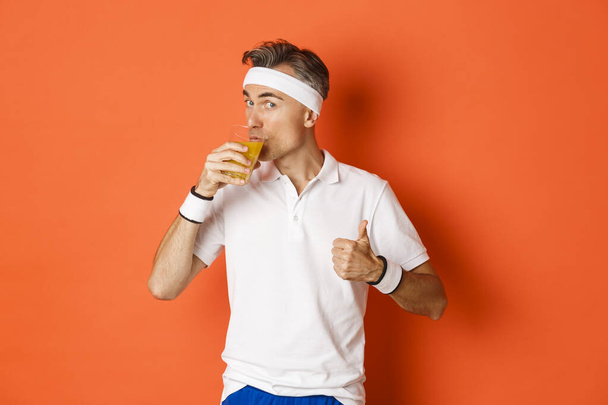 Concepto de entrenamiento, gimnasio y estilo de vida. Retrato del guapo chico de mediana edad, mostrando los pulgares hacia arriba y bebiendo jugo, de pie sobre fondo naranja - Foto, imagen