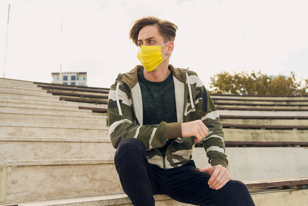 Spor kıyafetli, tıbbi maske takan genç bir adamın portresi. Covid-19 ölçümlerinden dolayı sarı tıbbi maske takan çekici bir erkek model. Beyaz adam kendini korumak için maske takıyor. - Fotoğraf, Görsel