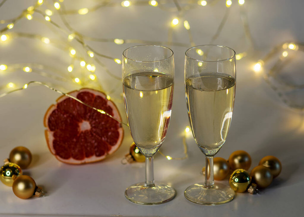 zwei Gläser Champagner, rosa Grapefruit, goldene Glaskugeln und verschiedene Weihnachtsdekorationen, beleuchteter Hintergrund einer Kette weißer Lichter, Weihnachtswartezeit, Winter - Foto, Bild
