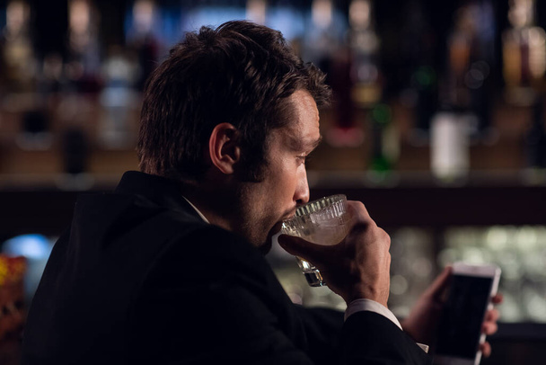 Молодой человек выпивает коктейль из бокала в баре, держа в руках смартфон, крупным планом - Фото, изображение