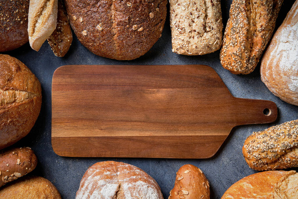 A variety breads surrounded by wooden cutting board on a dark surface. Top view, copy space. Food bakery shop concept banner background. - Fotografie, Obrázek