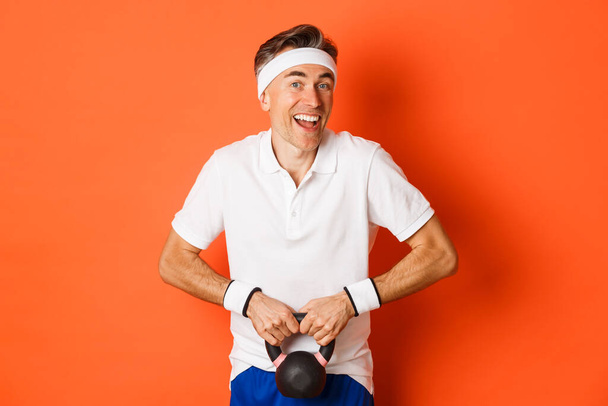 Koncept cvičení, tělocvičny a životního stylu. Obrázek pohledného chlapa středního věku, jak dělá sportovní cvičení, zvedá kettlebell a usmívá se, stojí nad oranžovým pozadím - Fotografie, Obrázek
