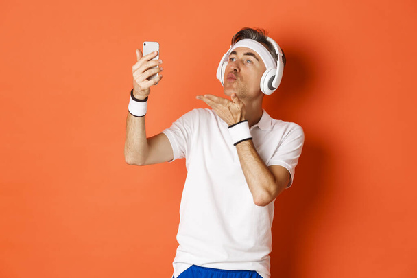 Portrait of attractive middle-aged man in gym uniform, wearing headphones, sending air kiss at mobile phone camera, taking selfie or having video call, standing over orange background - Photo, Image