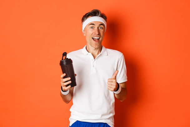 Porträt eines gesunden und aktiven Fitness-Typen mittleren Alters, der Turnuniform trägt, Daumen nach oben zeigt, Wasser trinkt und vor orangefarbenem Hintergrund steht - Foto, Bild
