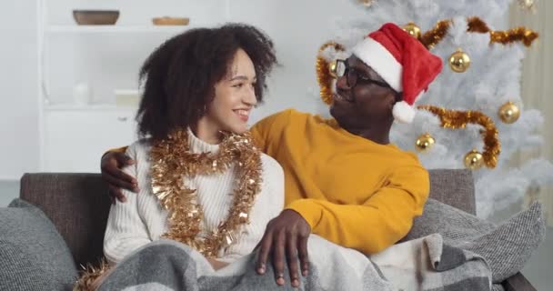 Afro-americano mulher esposa namorada encaracolado menina e homem negro namorado marido sentado juntos no sofá na sala de estar com decoração de Ano Novo no fundo da árvore de Natal ternamente abraçando - Filmagem, Vídeo