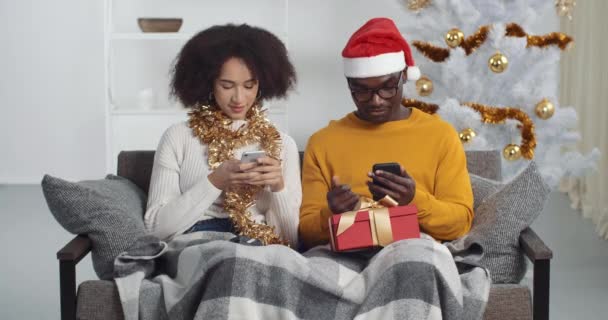 Afro-americanos pareja de amigos sentados en el sofá en trajes festivos mirando a los teléfonos inteligentes móviles de navegación de chat en internet chico mostrando chica vídeo musical novia bailando se mueve cabeza a la música - Metraje, vídeo