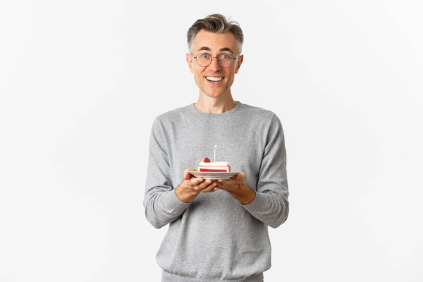 Wizerunek przystojnego mężczyzny w średnim wieku wyglądającego na zaskoczonego i szczęśliwego, któremu gratulujemy urodzin, trzymającego tort b-day i uśmiechającego się zdumionego, świętującego na białym tle - Zdjęcie, obraz