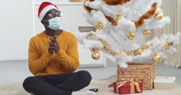 Afrikanischer schwarzer Mann im Neujahrsanzug sitzt in der Nähe des Weihnachtsbaums und trägt eine medizinische Maske auf dem Gesicht, gratuliert zum Urlaub aus der Ferne und redet vor laufender Kamera Daumen hoch, als würde er zum Abschied winken - Filmmaterial, Video