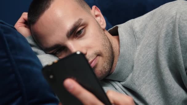 Junger Kaukasier nutzt Smartphone, während er zu Hause auf dem Sofa sitzt Mann surft im Internet, schaut sich Videos an und nutzt soziale Netzwerke zu Hause. - Filmmaterial, Video