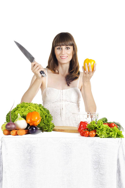 Piękna młoda dziewczyna przygotowuje sałatkę przy stole ze świeżymi warzywami na białym, odosobnionym tle. Trzyma bakłażana w dłoni.. - Zdjęcie, obraz