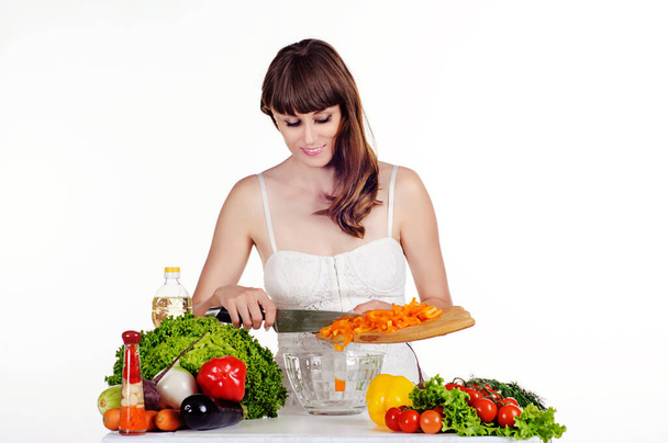 Hermosa joven prepara una ensalada en la mesa con verduras frescas sobre un fondo blanco aislado. La chica pone la pimienta en un plato transparente - Foto, imagen