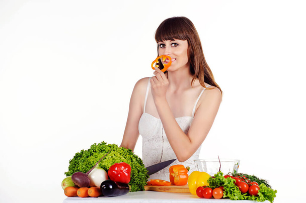 Hermosa joven prepara una ensalada en la mesa con verduras frescas sobre un fondo blanco aislado. La chica sostiene un anillo de pimienta naranja - Foto, imagen
