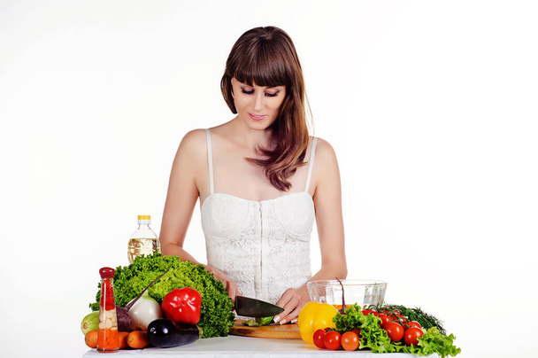 Красивая молодая девушка готовит салат за столом со свежими овощами на белом изолированном фоне. Девушка сдвигает оругци в прозрачной тарелке - Фото, изображение