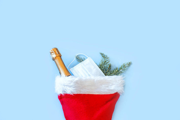 Red Santa hat, spruce branches and bottle champagne on blue background. Christmas and New Year during pandemic of coronavirus infection. Flat lay, top view, copy space. - Photo, Image