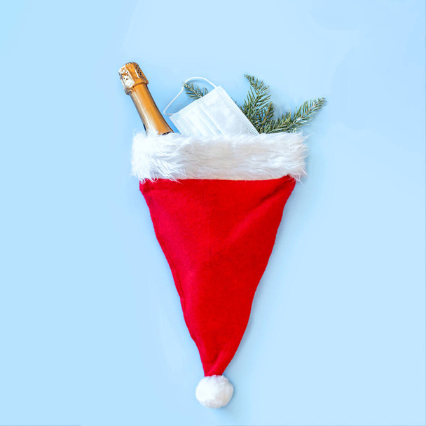 赤いサンタの帽子、青い背景にトウヒの枝とボトルシャンパン。コロナウイルス感染のパンデミックの間のクリスマスと新年。フラットレイアウト、トップビュー、コピースペース. - 写真・画像