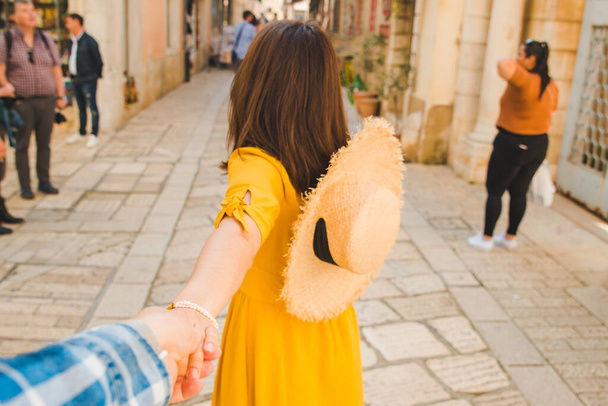 Beni izle konsept. Hasır şapkalı, sarı elbiseli bir kadın küçük bir tatil köyü caddesinde erkek eli tutarak yürüyordu. yaz tatili - Fotoğraf, Görsel