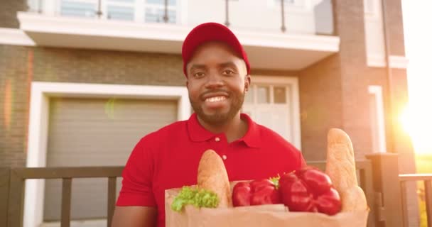 Retrato del joven mensajero afroamericano en uniforme rojo y sombrero parado en casa en la calle y sosteniendo el paquete con comida saludable. Trabajador de reparto masculino en uniforme con bolsa de productos sonriendo. - Imágenes, Vídeo