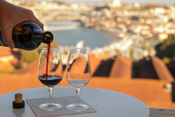 Pouring of fortified dessert ruby, tawny port wines in glasses with view on Douro river, porto lodges of Vila Nova de Gaia and city of Porto, Portugal, on sunset - Photo, Image