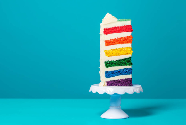 Single slice of birthday cake on a white cakestand minimalist on a blue background. Homemade cake with multicolored layers and buttercream - Photo, Image