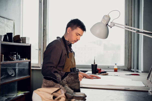 Un zapatero joven hace un dibujo para un patrón de zapatos de cuero en una mesa en su taller - Foto, Imagen