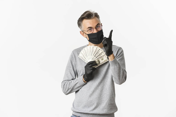 Concept van covid-19, sociale afstand en levensstijl. Portret van een gelukkige man van middelbare leeftijd in medisch masker, handschoenen en bril, met geld en wijzende vinger omhoog, glimlachend blij - Foto, afbeelding