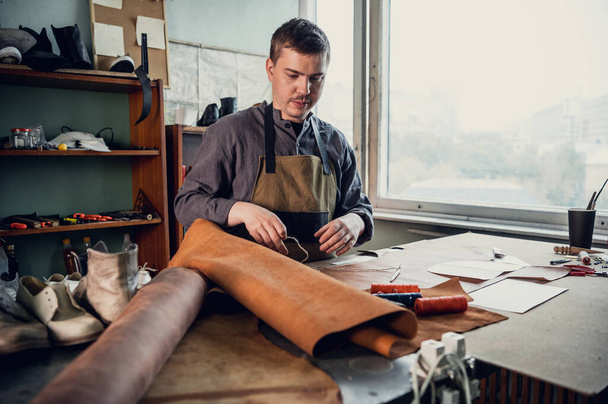 Un joven aprendiz en un taller de botas prepara el cuero para su uso posterior en una mesa grande - Foto, Imagen
