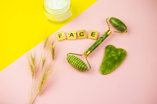 Grüne Gua Sha Gesichtsmassagegeräte. Walze aus grüner Quarzjade auf rosa-gelbem Hintergrund. Glas Sahne, Schriftzug Gesicht in Holzbuchstaben. Nahaufnahme, Draufsicht - Foto, Bild