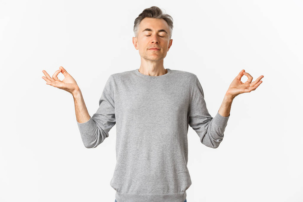 Portrait of calm and relaxed middle-aged man, breathing air freely, meditating with eyes closed and hands spread sideways, practice yoga over white background - Photo, Image