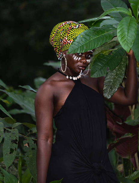 Африканская женщина, стоящая в настоящих джунглях с племенными отметинами на лице и африканскими цветами в качестве головного убора, фото сделано в Гане - Фото, изображение