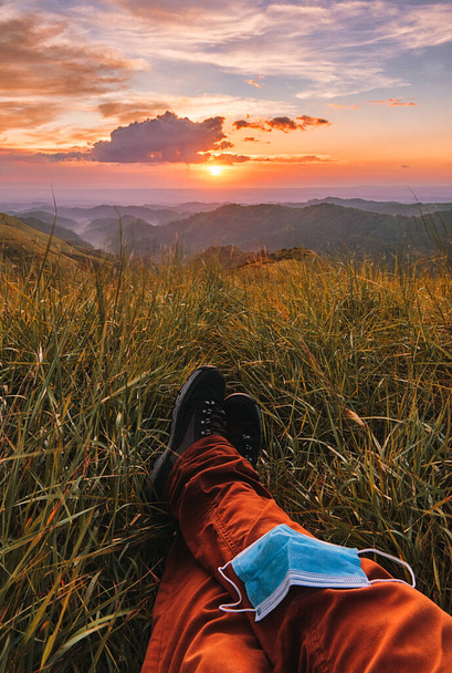 Plan vertical des pieds d'une personne couchés sur le sol avec masque chirurgical au large entouré de collines lors d'un beau coucher de soleil - Photo, image