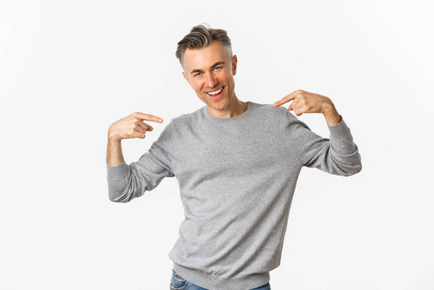 Image of handsome and confident middle-aged man with short grey hairstyle, pointing at himself and smiling, show-off over white background - Photo, Image