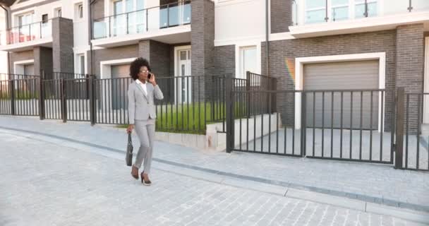 Afroamericano joven elegante hermosa agente de bienes raíces mujer caminando por la calle en el barrio suborbe y hablando por teléfono móvil. Mujer de negocios hablando por teléfono celular al aire libre en las afueras. - Imágenes, Vídeo
