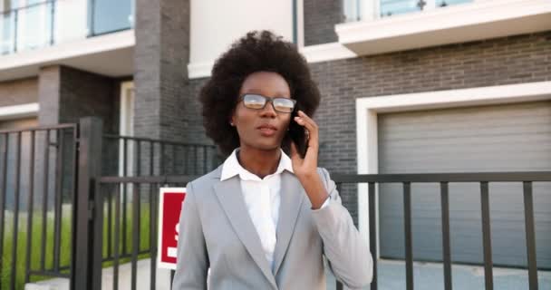 Afroamericano joven mujer elegante agente de bienes raíces en gafas en el barrio periférico y hablando por teléfono móvil. Mujer de negocios buscando casa y hablando por celular. Vivienda en venta. - Imágenes, Vídeo