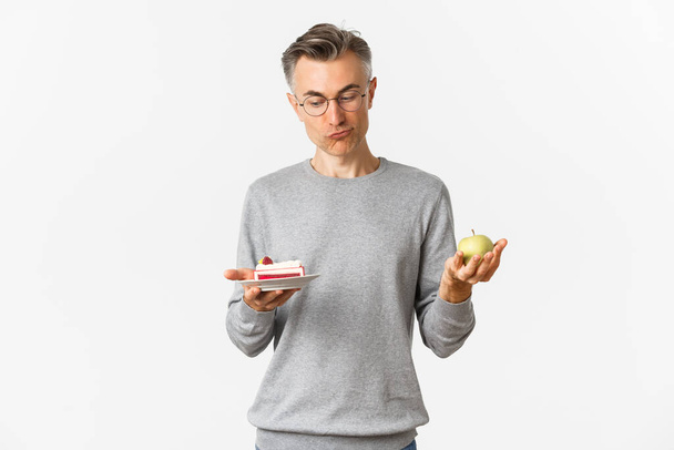 Portrait of thoughtful middle-aged man in glasses, making decision between tasty cake and green apple, standing over white background - Photo, Image