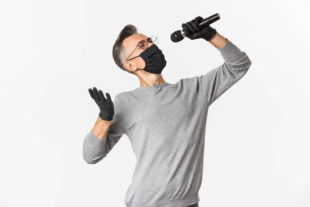Koronaviruksen, elämäntavan ja karanteenin käsite. Muotokuva houkutteleva keski-ikäinen mies musta lääketieteellinen naamio ja käsineet, laulaa mikrofonissa, seisoo valkoinen tausta - Valokuva, kuva