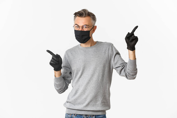 Concept of coronavirus, lifestyle and quarantine. Image of middle-aged man with grey hair, wearing black medical mask, gloves and glasses, pointing fingers sideways, showing copy space - Photo, Image