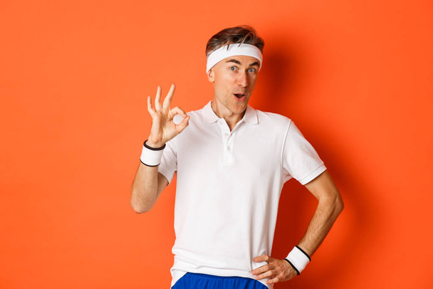 Pojetí sportu, fitness a životního stylu. Portrét ohromeného dospělého muže v pracovní uniformě, ukazuje v pořádku znamení a vypadá ohromeně, stojící nad oranžovým pozadím - Fotografie, Obrázek