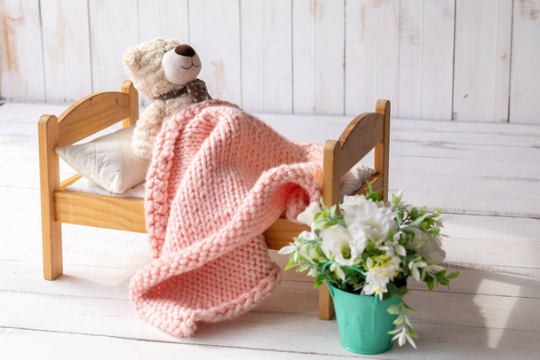Um urso de pelúcia está deitado em uma pequena cama de madeira, coberta com um cobertor de malha. Próximo a ele é um pote com uma flor artificial. Conceito de sono do bebê - Foto, Imagem