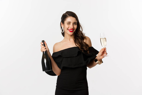 Feest- en feestconcept. Stijlvolle brunette vrouw in glamour jurk holding fles en glas champagne, plezier hebben op nieuwjaarsvakantie - Foto, afbeelding