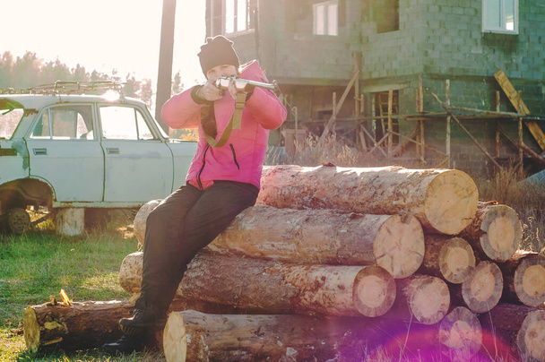 Una chica dispara un arma mientras está sentada en un tronco torcido contra una casa abandonada - Foto, Imagen