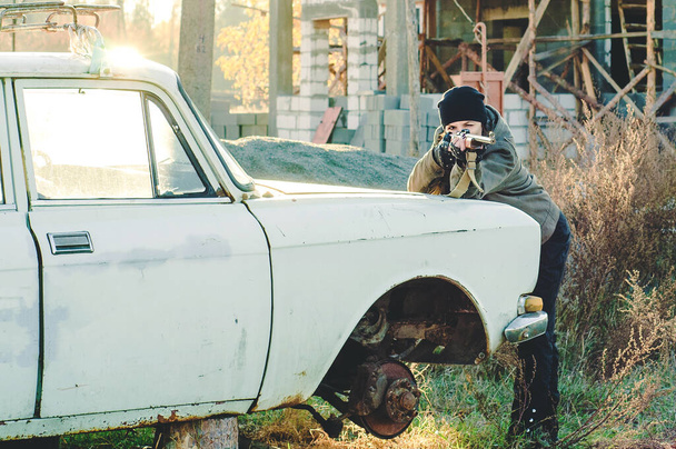 A girl shoots a gun, leaning on a broken car against an abandoned home - Foto, imagen