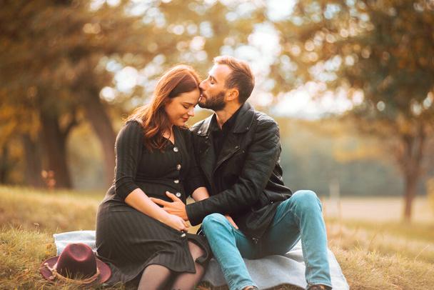Photo of happy pregnant couple sitting on the banch in park, beautiful forehead kiss - Photo, Image