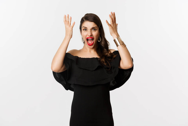 Fashion and beauty. Angry woman in black dress, shouting mad and shaking hands, grimacing outraged at camera, standing over white background - Photo, Image