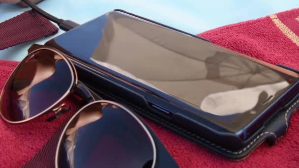 Riflessi sfocati negli occhiali da sole e sullo schermo dello smartphone in cima all'asciugamano rosso in spiaggia - Filmati, video