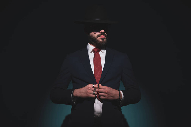 guay hombre de negocios guapo cerrando su chaqueta, mirando hacia otro lado y usando un sombrero negro en una pose de moda - Foto, imagen